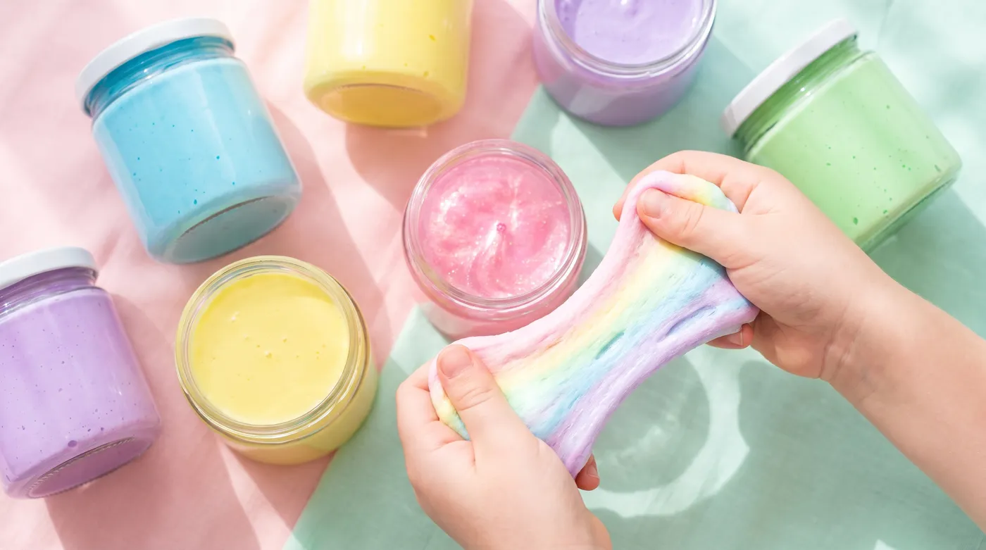 Pots de slime colorés bien fermés sur fond pastel, avec des mains qui étirent un slime brillant